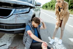 A woman looks on as an injured man sits by a car with visible damage.
