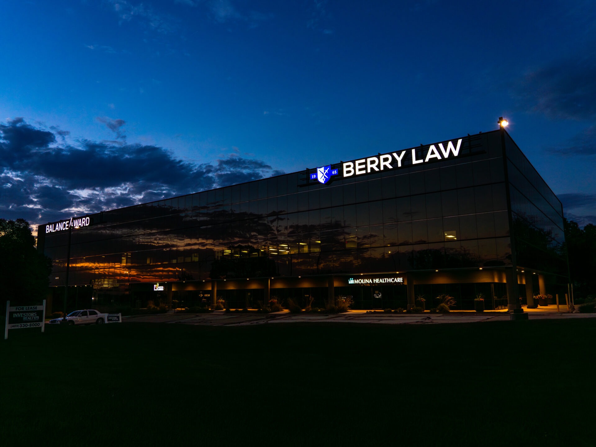 Exterior of Berry Law’s new West Omaha office illuminated at night, featuring modern architecture, large glass windows, and the Berry Law shield logo glowing above the entrance.